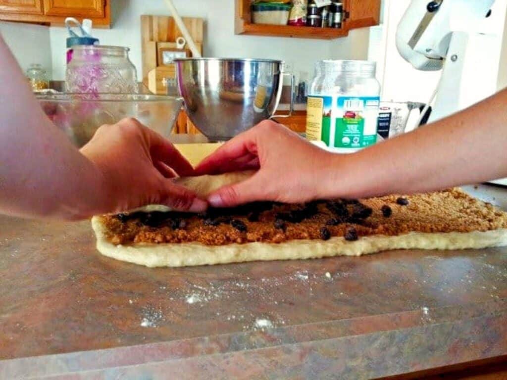 Two hands roll up no yeast cinnamon roll dough filled with brown sugar and raisins on a kitchen counter, with baking supplies visible in the background.