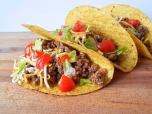 Three hard-shell honey habanero tacos filled with seasoned ground beef, shredded cheese, lettuce, and diced tomatoes on a wooden surface.