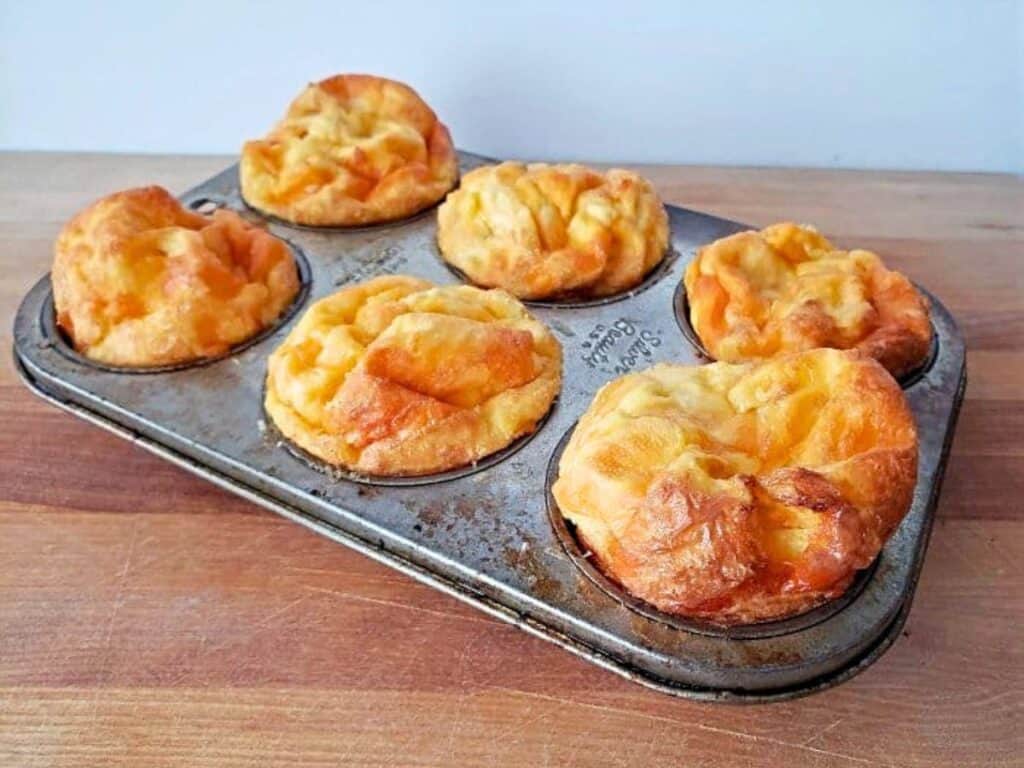 A metal muffin tin holding six baked popovers sits on a wooden surface against a plain light background.