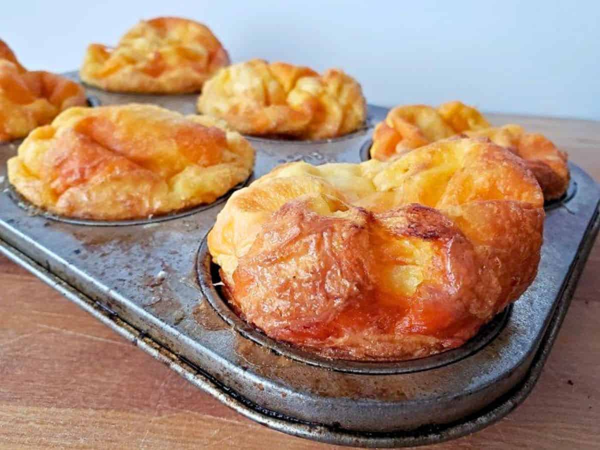 A muffin tin filled with six golden brown, baked popovers sits on a wooden surface against a plain background.