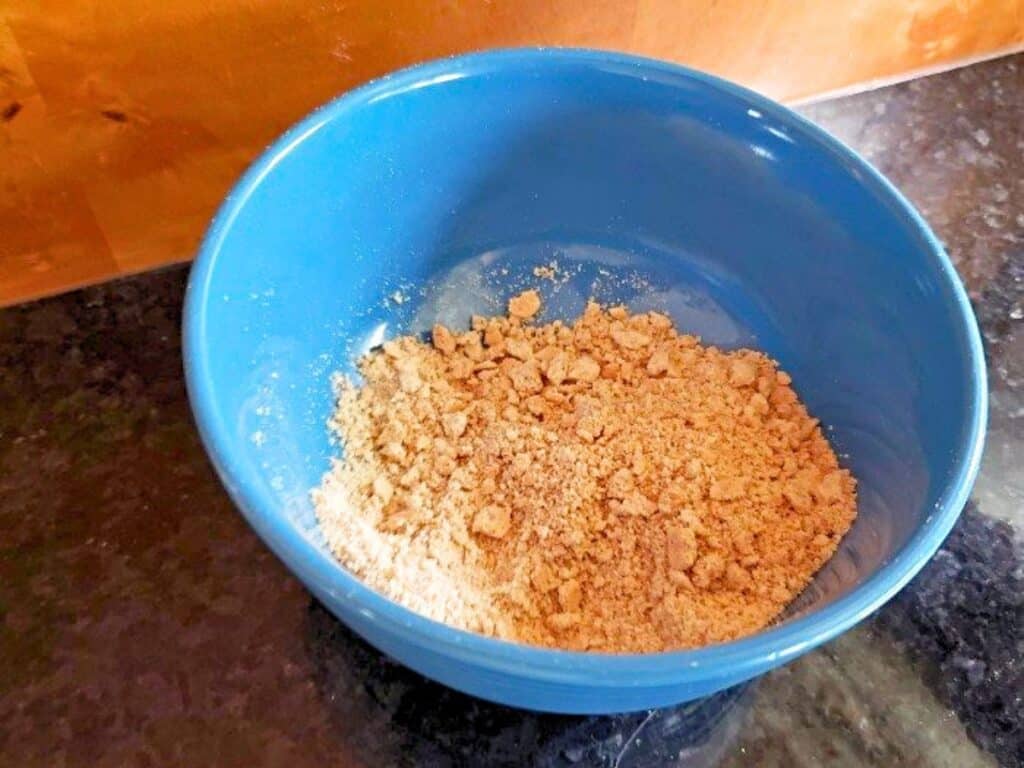 Blue bowl containing a mixture of crushed biscuit crumbs and edible s&rsquo;mores cookie dough on a dark countertop with a wooden background.