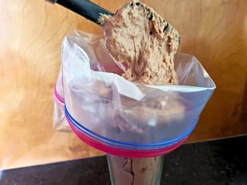 A plastic bag in a glass is being filled with brown batter using a spatula, likely to be used as a piping bag.