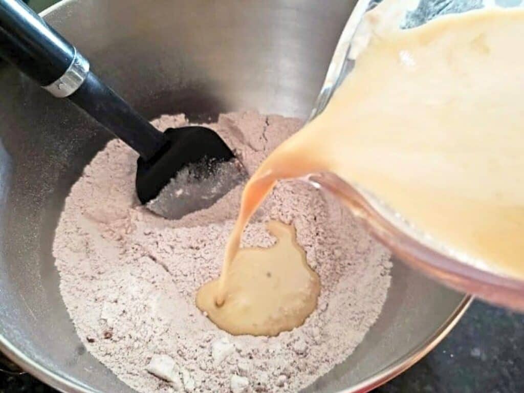 A spatula rests in a mixing bowl with dry ingredients as a liquid mixture is poured in from a measuring cup.