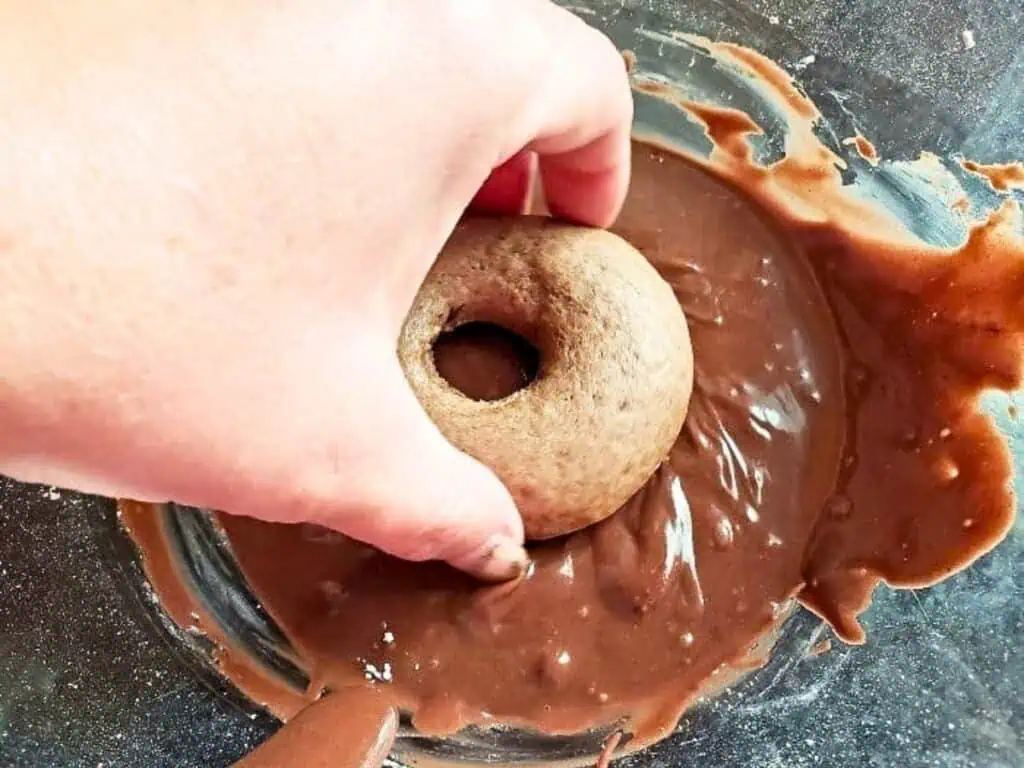 A hand dipping a baked donut into a bowl of chocolate glaze.