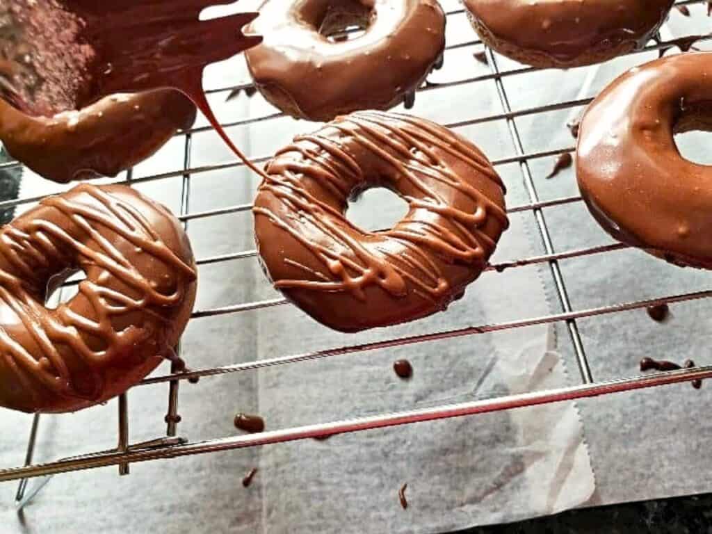 Chocolate-covered donuts resting on a metal cooling rack with parchment paper underneath; some donuts are being drizzled with more chocolate.