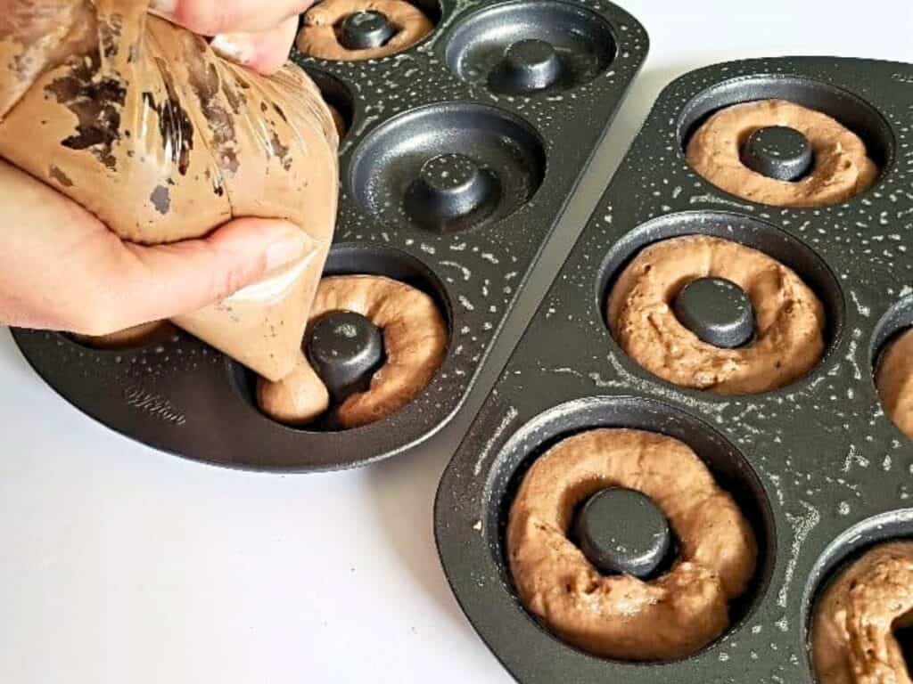 Hands pipe chocolate batter into a greased donut pan with several cavities already filled, preparing to bake homemade donuts.