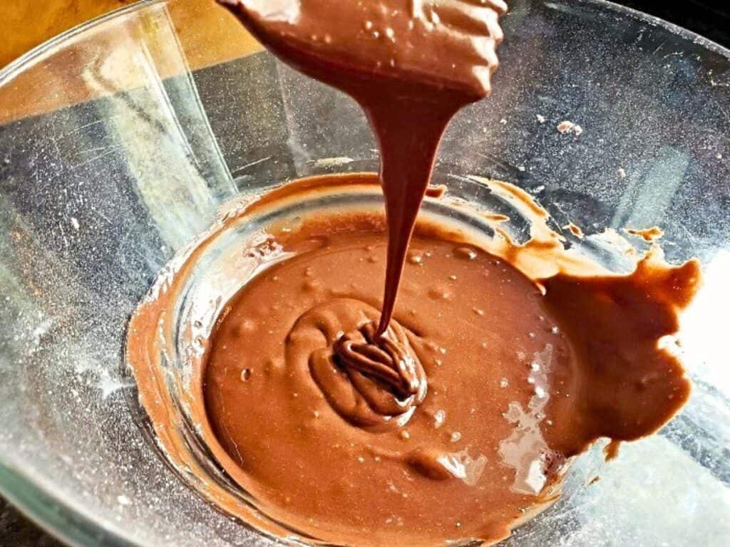 A glass bowl contains chocolate cake batter being poured from a spatula, with swirls visible on the surface.