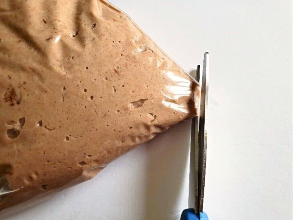 A pair of scissors cuts the corner of a plastic bag filled with light brown batter or mixture, set against a white background.