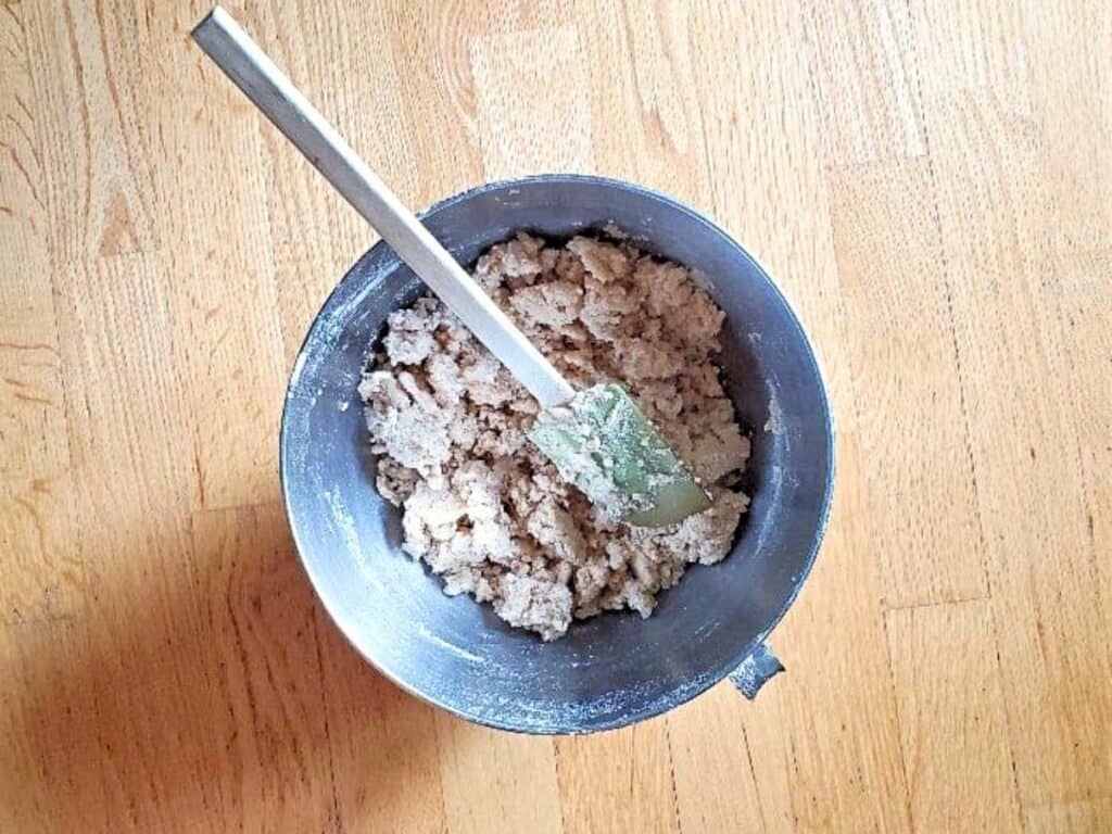 A metal mixing bowl with cookie dough for cherry crumb bars and a spatula sits on a wooden surface.