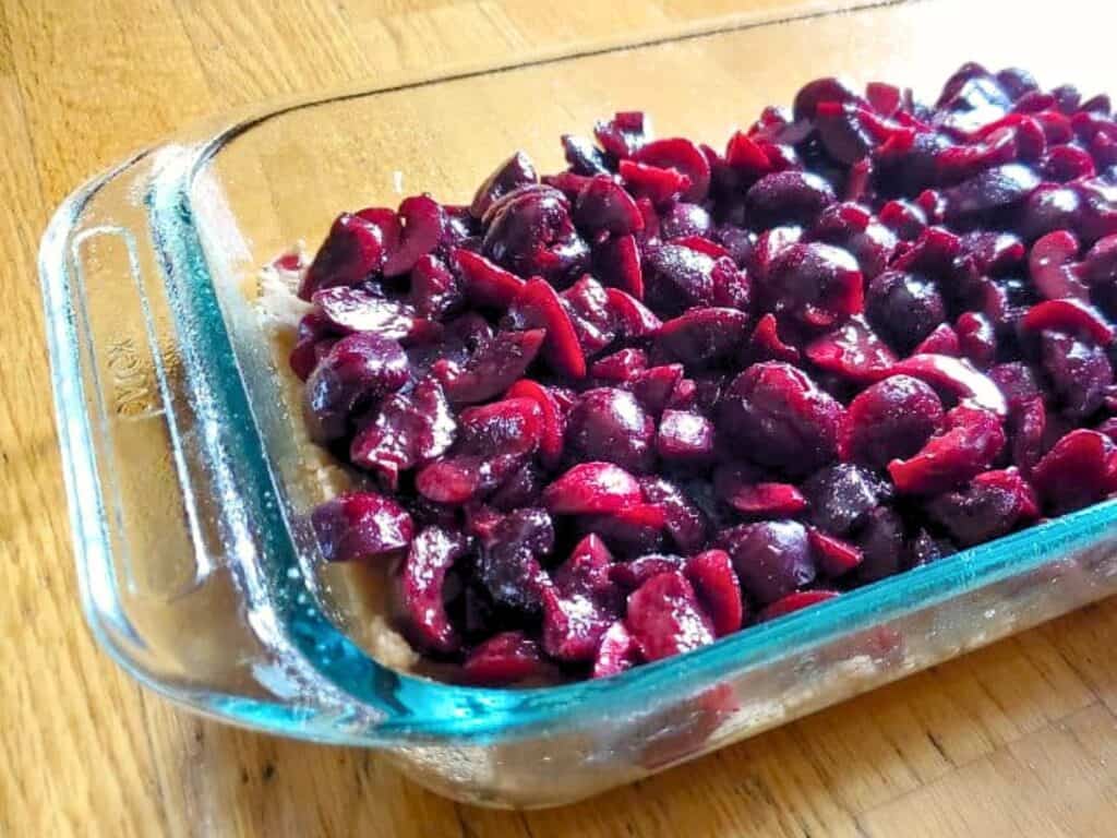 A glass baking dish filled with chopped dark cherries spread over a crust layer, creating delicious cherry crumb bars, is placed on a wooden surface.
