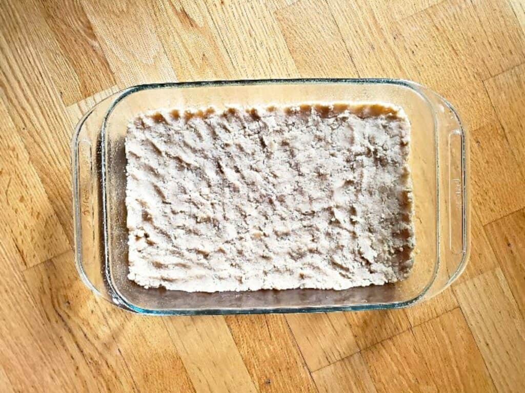 A glass baking dish filled with unbaked or partially baked cherry crumb bars, featuring a crumbly, textured topping, sits on a wooden surface.