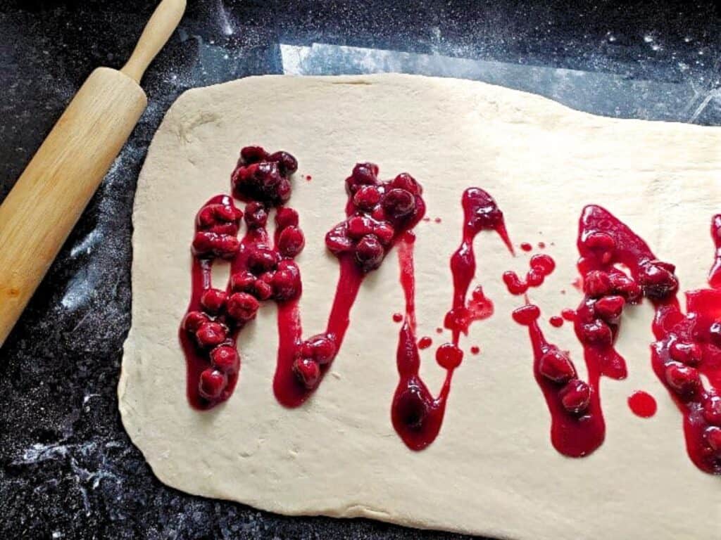 Rolled dough on a floured surface with cherry filling spread on top, ready to become delicious cherry cinnamon rolls; a wooden rolling pin lies nearby.