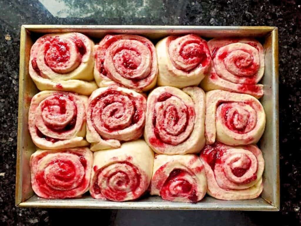 A rectangular baking pan filled with twelve unbaked strawberry swirl rolls, reminiscent of cherry cinnamon rolls, is shown from above on a dark countertop.