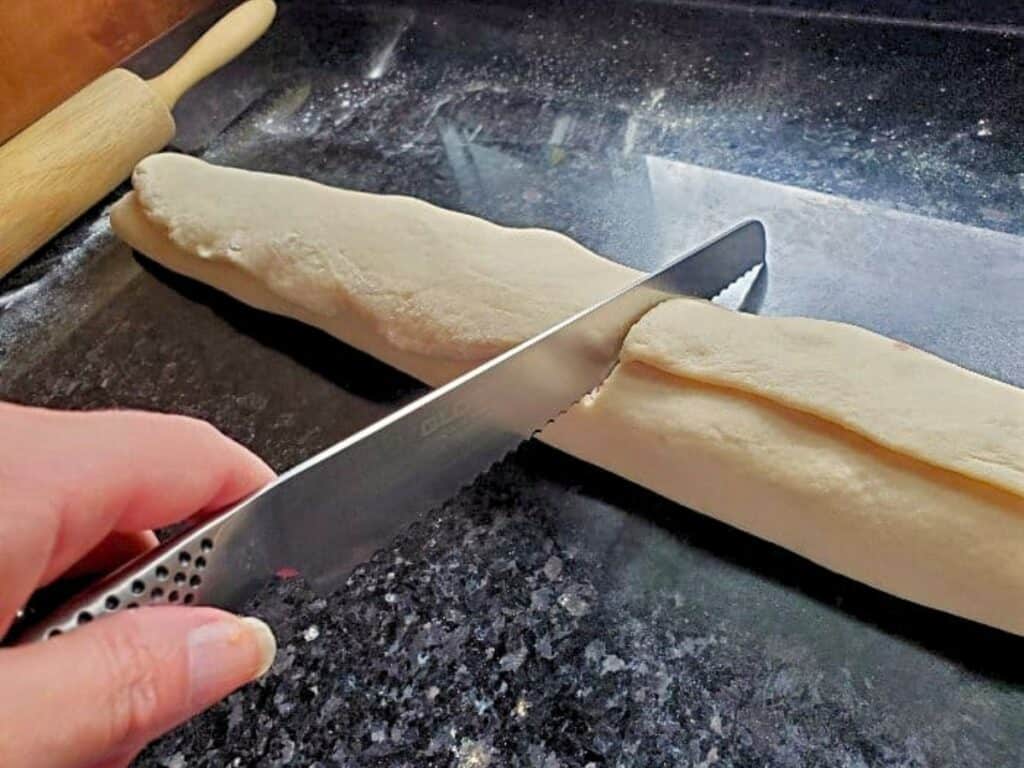 A hand uses a serrated knife to cut a long piece of dough for cherry cinnamon rolls on a dark countertop, with a rolling pin visible in the background.