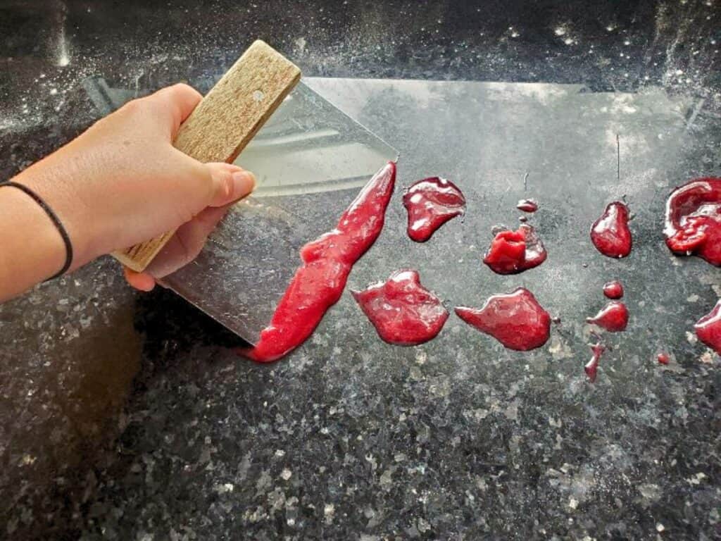 A hand uses a bench scraper to gather small piles of cherry cinnamon rolls dough or batter on a black granite countertop.