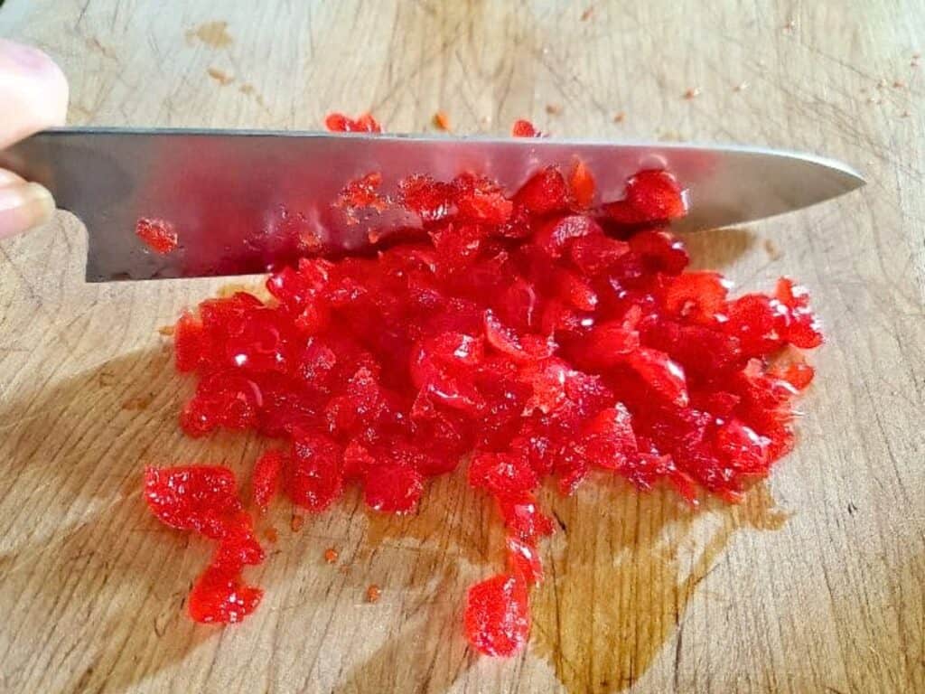 A hand holds a knife chopping bright red candied cherries on a wooden cutting board.