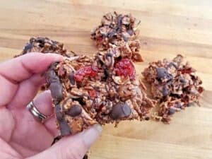 A hand holds a chocolate-drizzled granola bar with visible pieces of oats and dried cherries. Three more bars are on a wooden surface in the background.