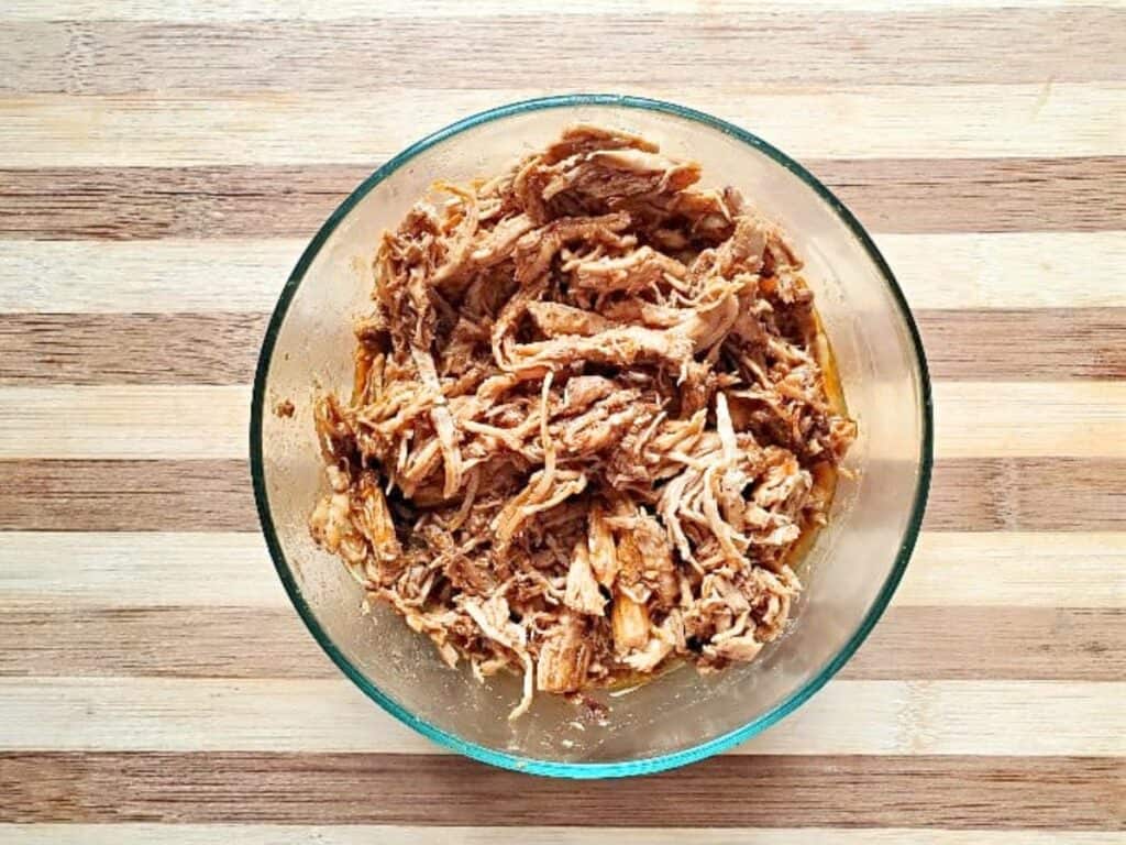 A clear glass bowl filled with Instant Pot BBQ shredded chicken sits on a light wooden surface with alternating light and dark stripes.
