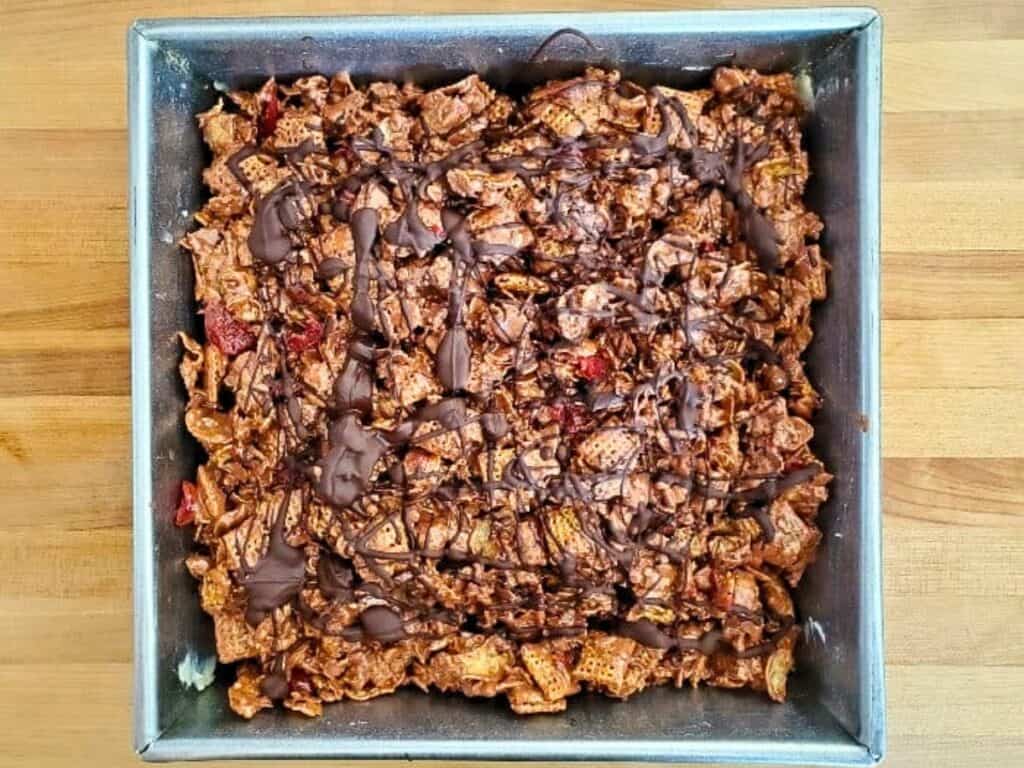 A square baking pan filled with a chocolate-drizzled cereal and nut mixture sits on a wooden surface.