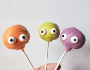 Image shows a hand holding 3 monster cake pops against a grey background.