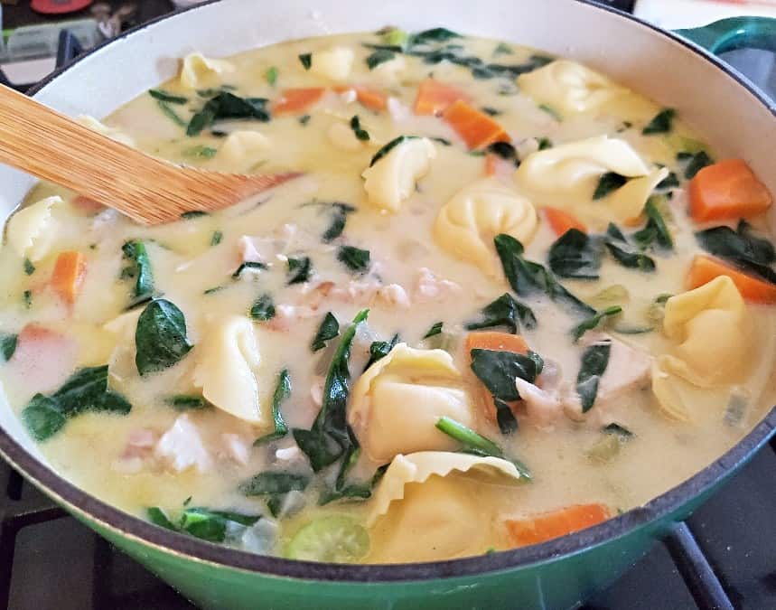 Image shows Chicken tortellini soup in a ceramic enameled pot with a wooden spoon stirring it.