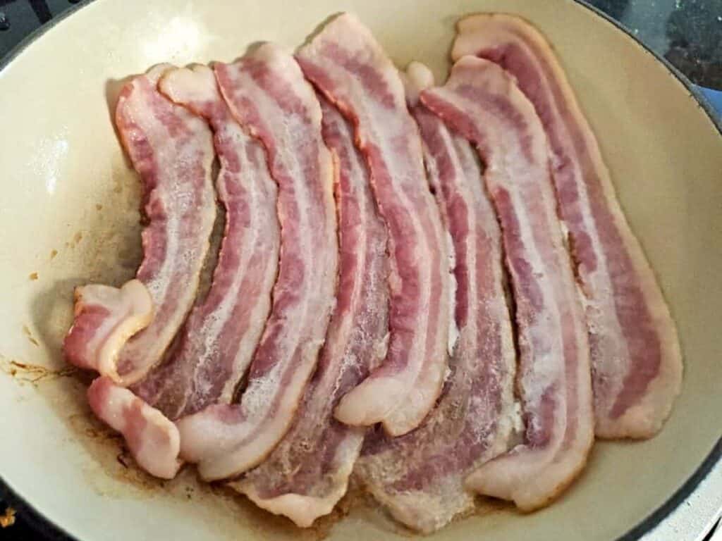 Six raw bacon strips are laid out side by side in a frying pan, beginning to cook—the perfect start for making a savory hot bacon cheddar dip.