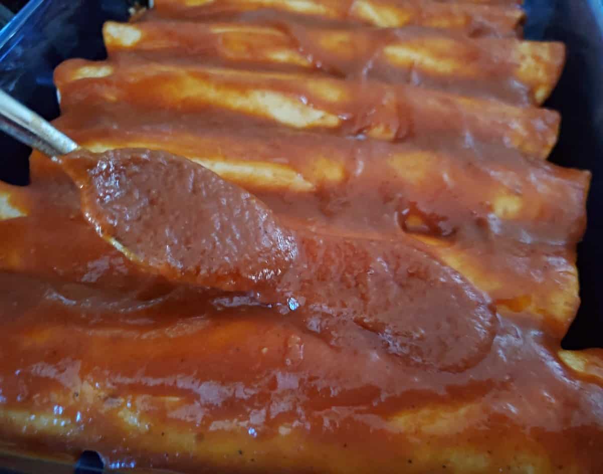 A wooden spoon has red enchilada sauce dripping off it into a blue pot filled with the remainder of the homemade enchilada sauce.