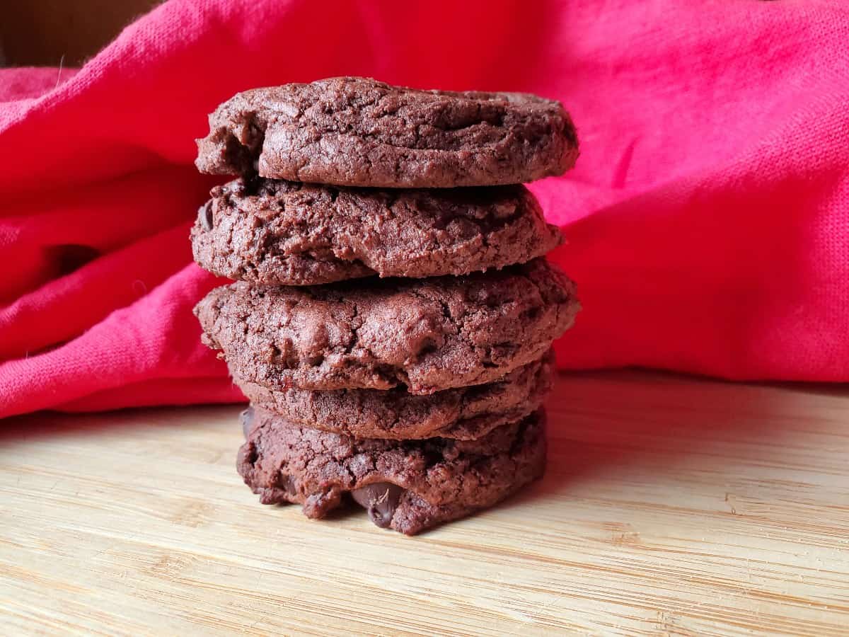 Image shows close-up of a hand holding a bitten chocolate cookie with more cookies in the background on a wooden surface.