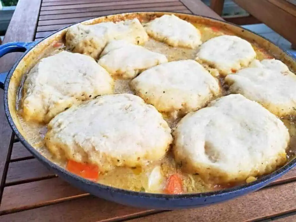 Image shows chicken stew with biscuits in a pot on a wooden table.