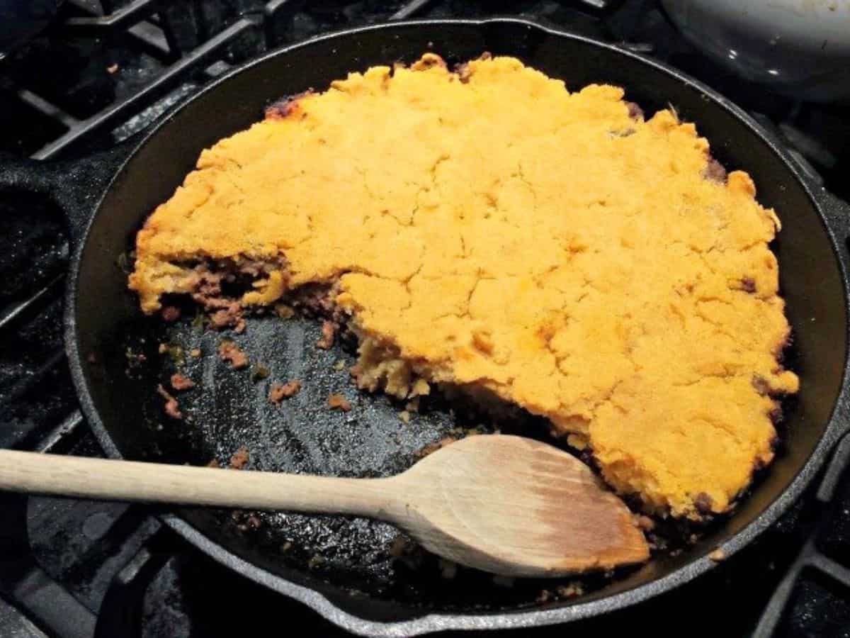 Image shows a partially served ground beef skillet tamales in a skillet with a wooden spoon.