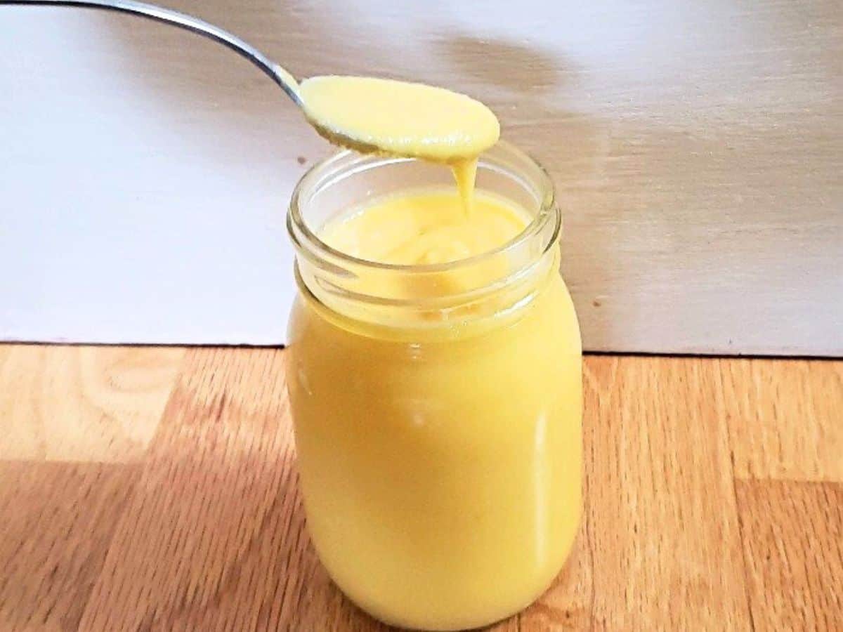 A front on photo shows mango curd in a glass jar with a spoon that has removed some curd.