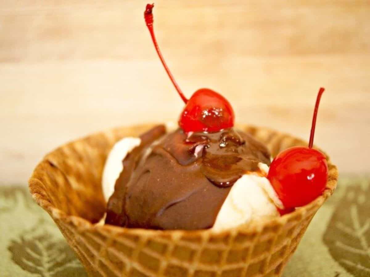 Image shows A waffle cone bowl containing vanilla ice cream topped with magic shell and two cherries.