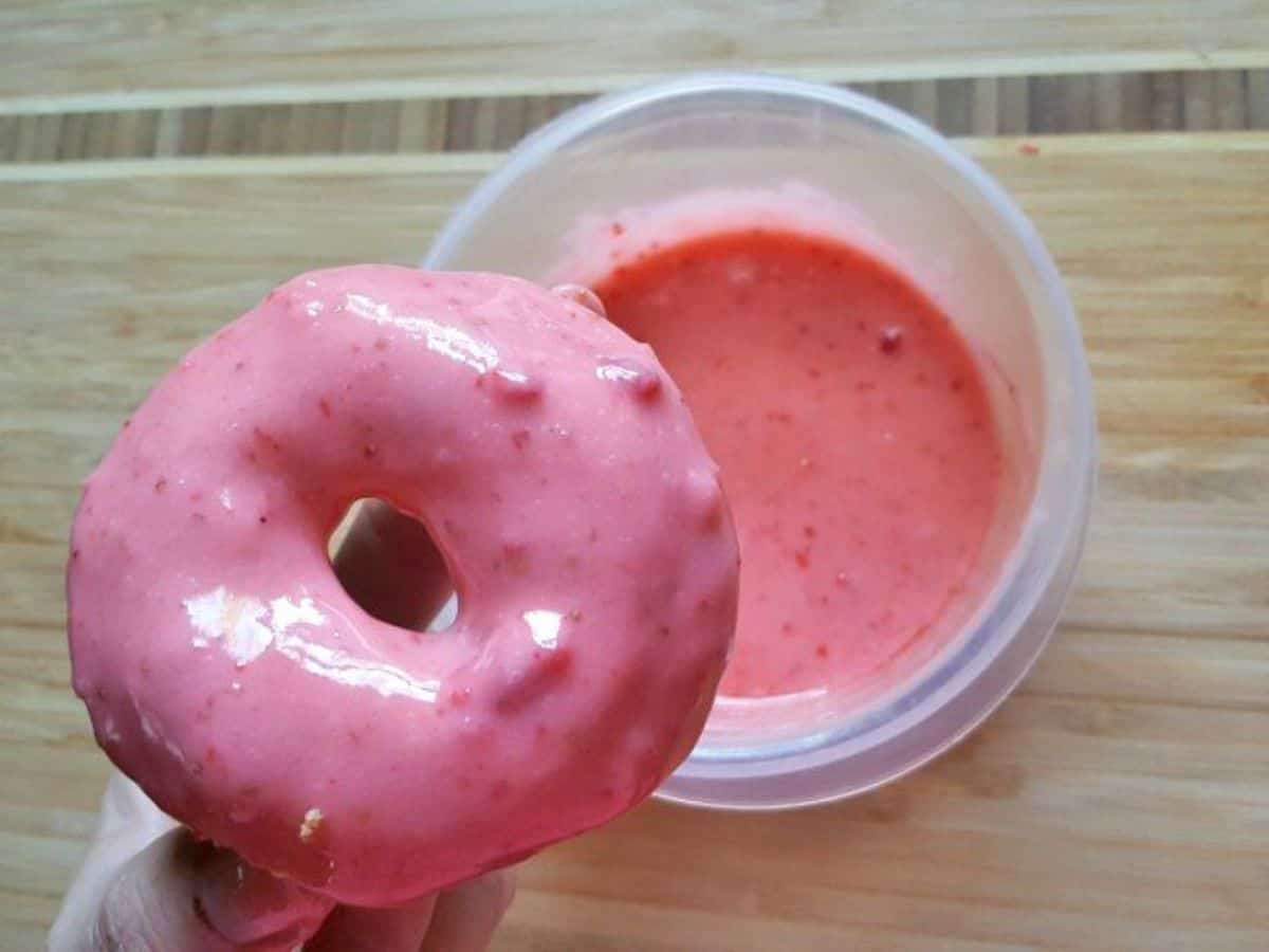Image shows a freshly dipped strawberry donut held over the glaze.