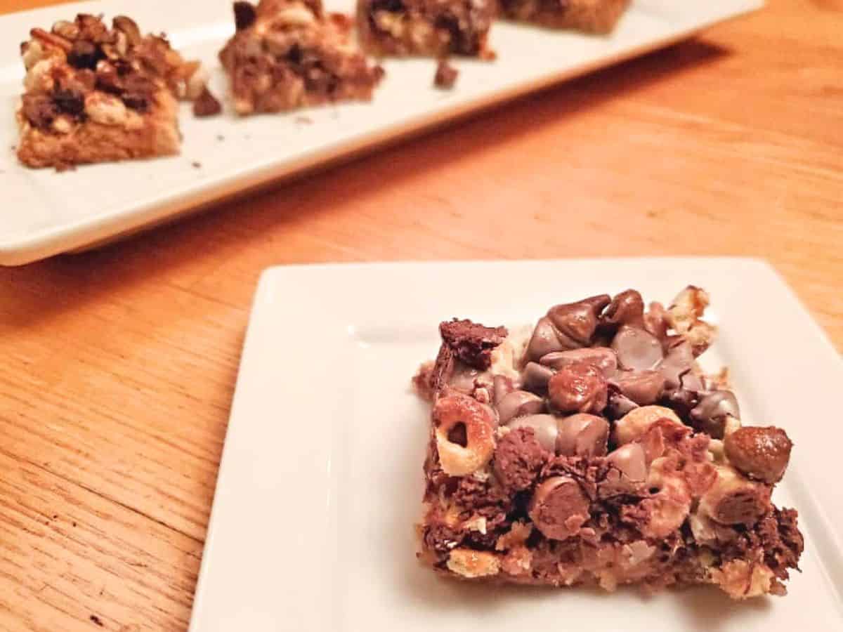 A dessert bar topped with chocolate chips and cereal pieces is placed on a white square plate on a wooden table. Another plate with a dessert bar is partially visible in the background.