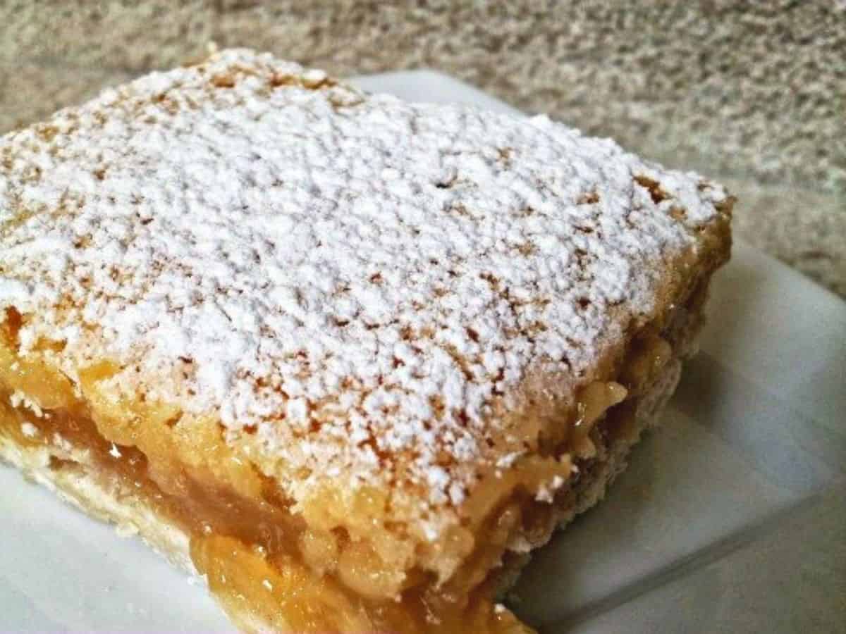 A close-up of a square slice of a lemon bar dessert, topped with a generous dusting of powdered sugar, served on a white plate.