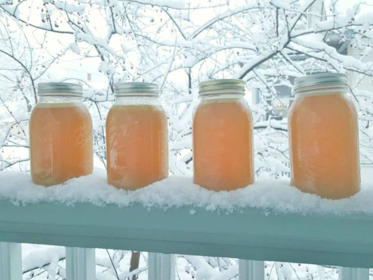 Image shows four mason jars of chicken stock sitting on a railing in the snow.