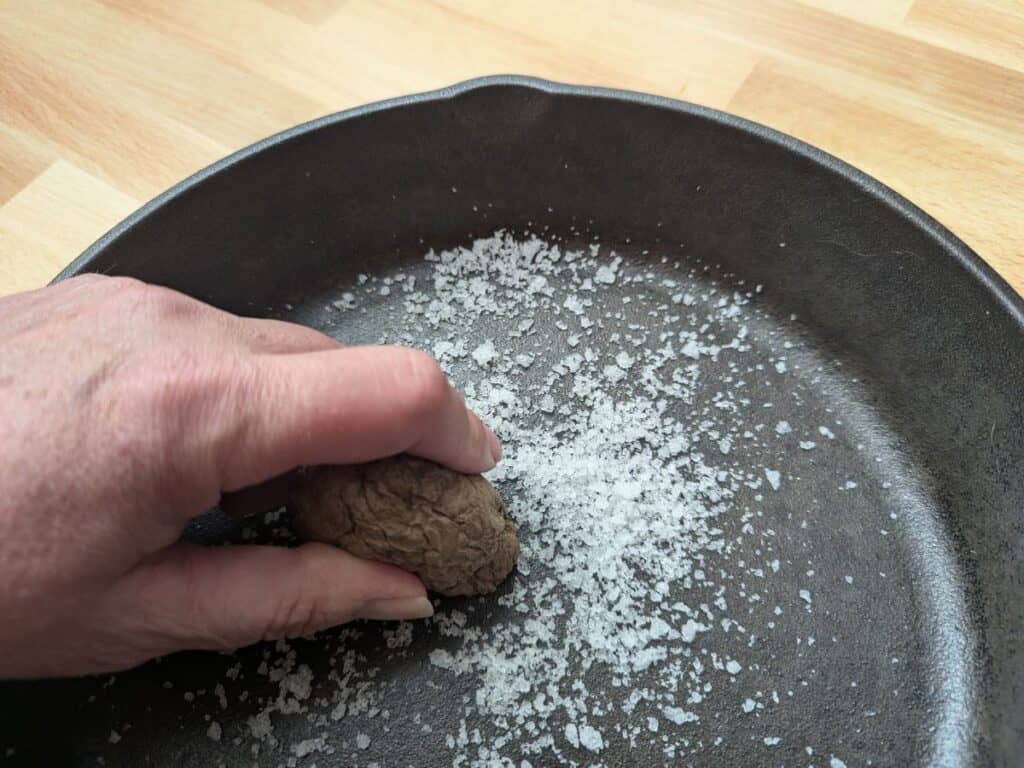 Image shows a Hand rubbing a potato and salt into a cast iron pan to clean it.