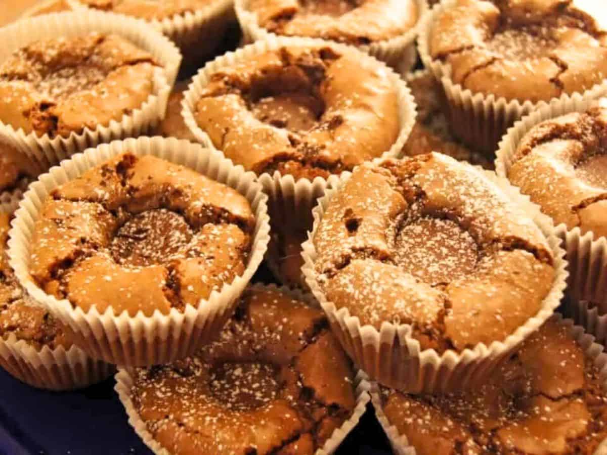 Image shows A close-up image of freshly baked brownies in muffin tins with a surprise in the middle and with a dusting of powdered sugar, placed in brown paper liners.