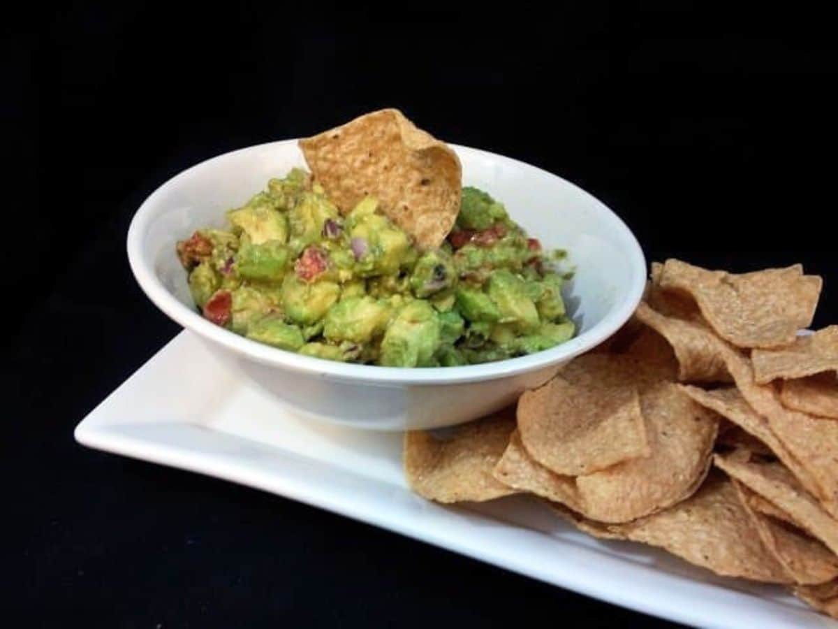Image shows a white bowl with guacamole and a chip sticking in it with more chips on a tray to the right of it.