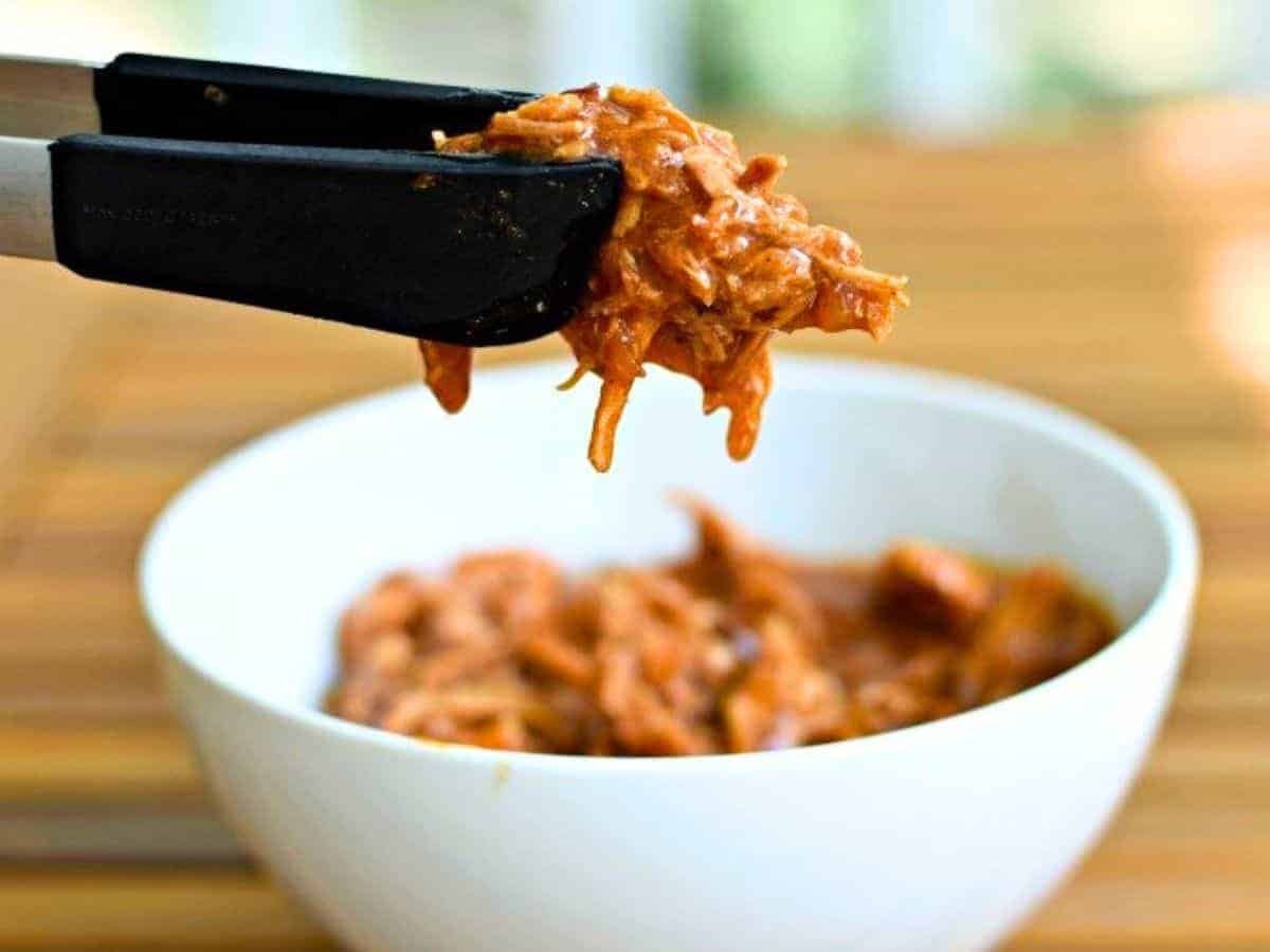 Tongs holding shredded barbeque pork over a white bowl with more pork inside.