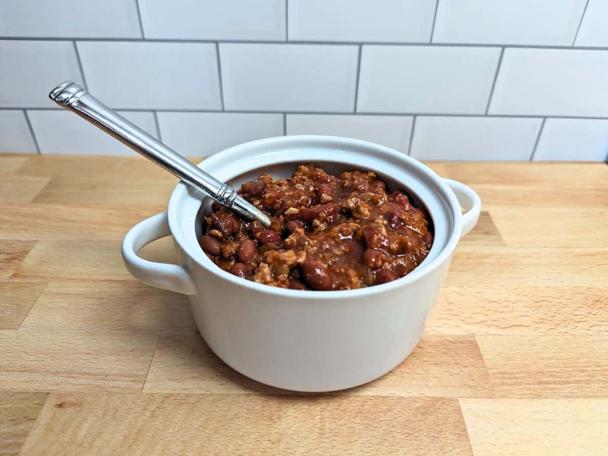 Image shows close-up of a spoonful of chili with beans, cheese, and corn, held above a pot filled with the same dish, placed on a wooden surface.