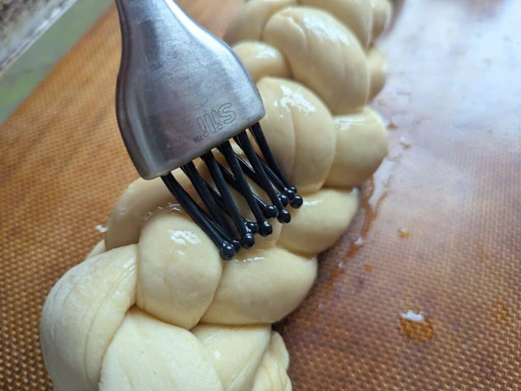 Image shows brushing egg wash on top of challah dough.