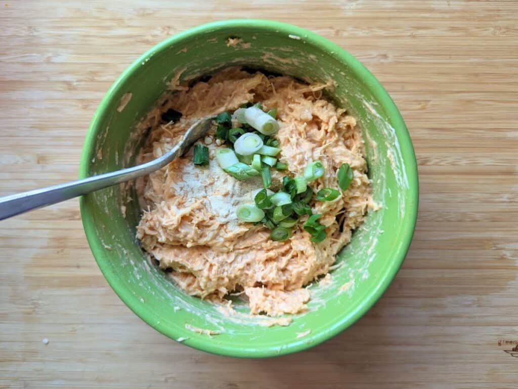Image shows buffalo chicken salad ingredients in a bowl, topped with sliced green onions and a sprinkle of garlic powder.