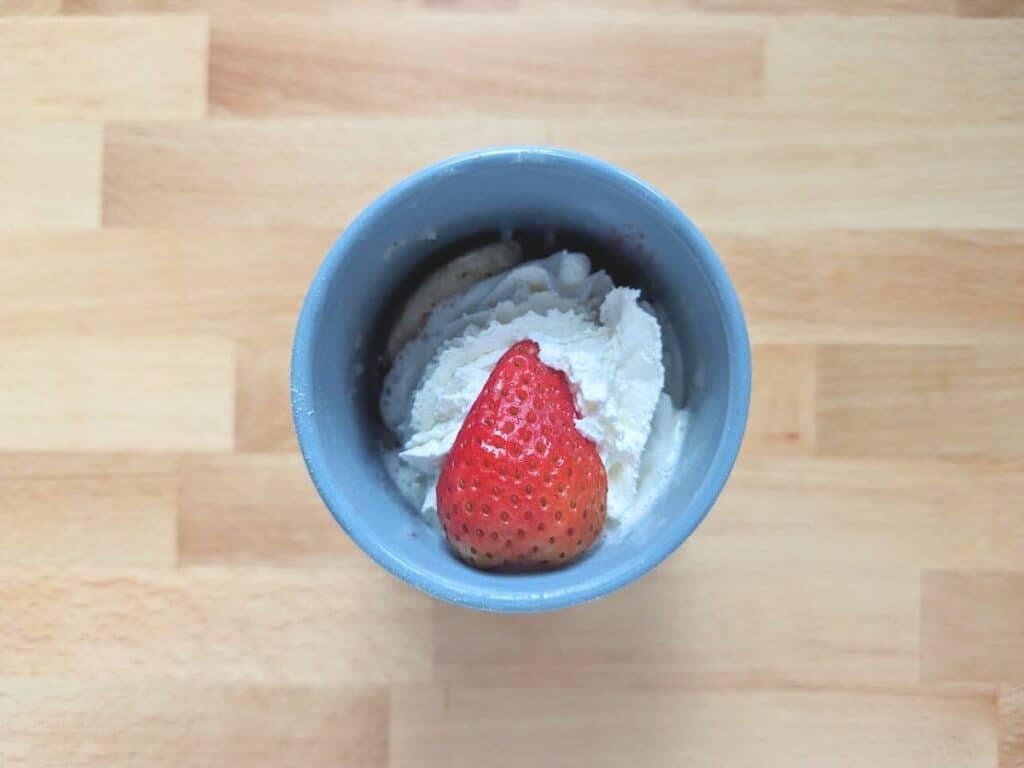 Image shows whipped cream and strawberries added on top of the cooked strawberry mug cake.