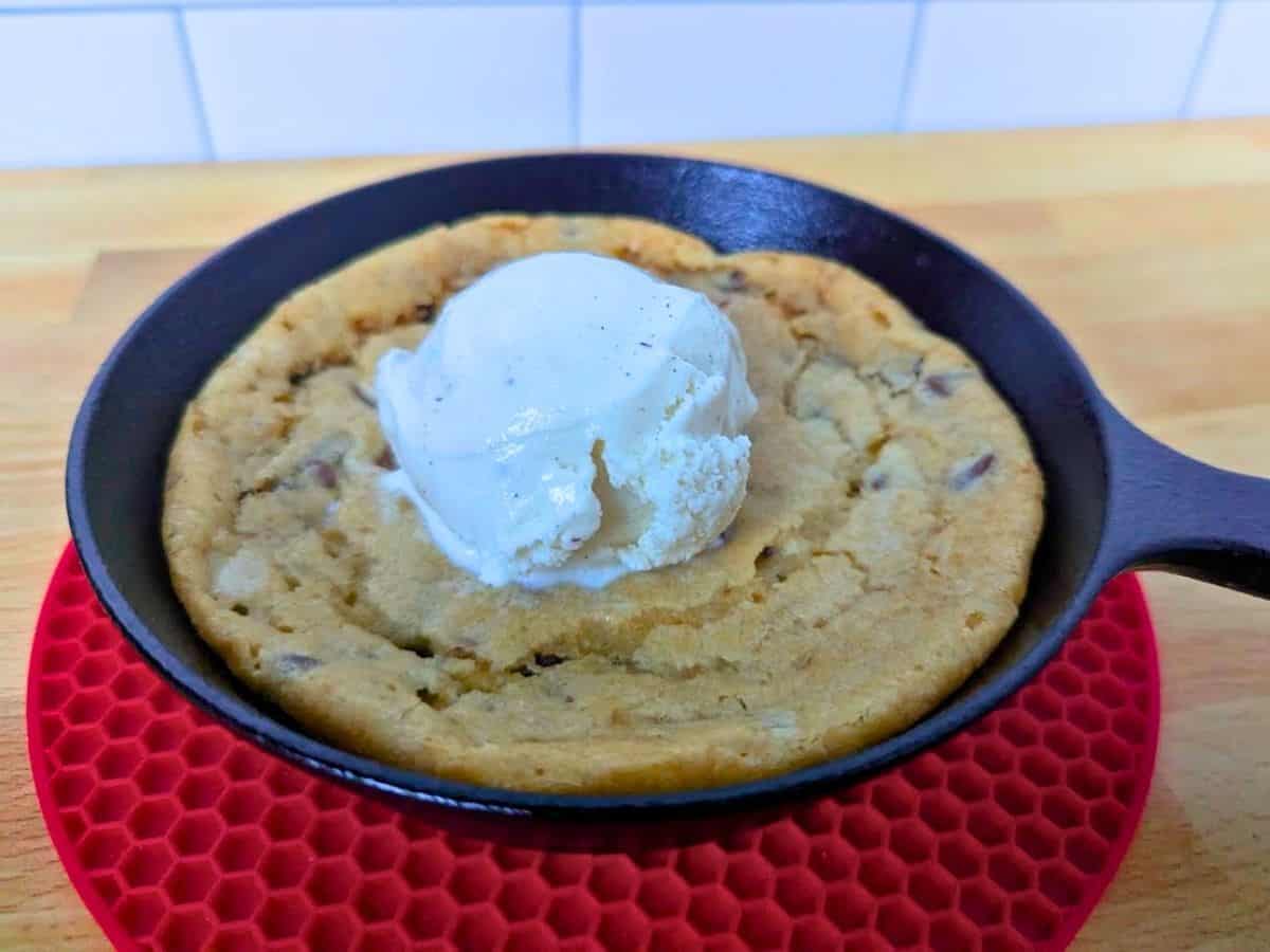 Image shows a chocolate chip cookie baked in a small cast iron skillet, topped with a scoop of vanilla ice cream, sits on a red silicone trivet on a wooden surface.