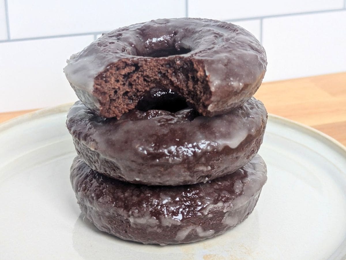 Image shows a stack of glazed chocolate donuts sits on a white plate atop a wooden surface, with a tiled wall in the background.