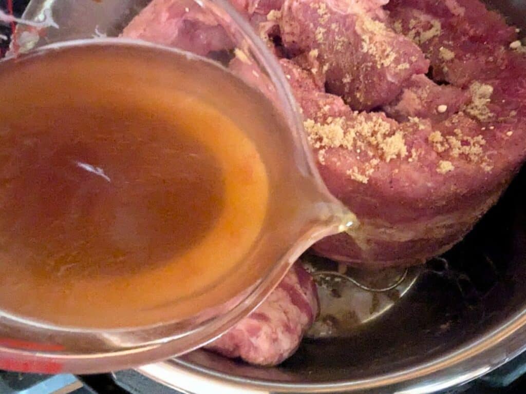 Image shows apple juice being poured over marinated pork ribs in a pot.