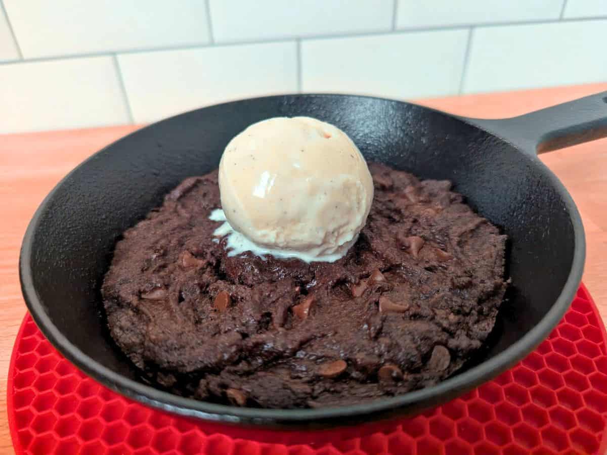 Image shows a chocolate chip skillet cookie topped with a scoop of vanilla ice cream, served in a black pan on a red mat.