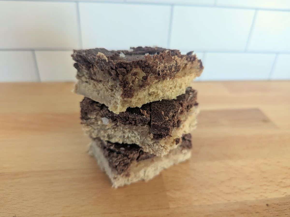 Image sjows a hand holds a partially bitten chocolate and caramel layered treat with a crumbly base, with more treats stacked in the background on a wooden surface.