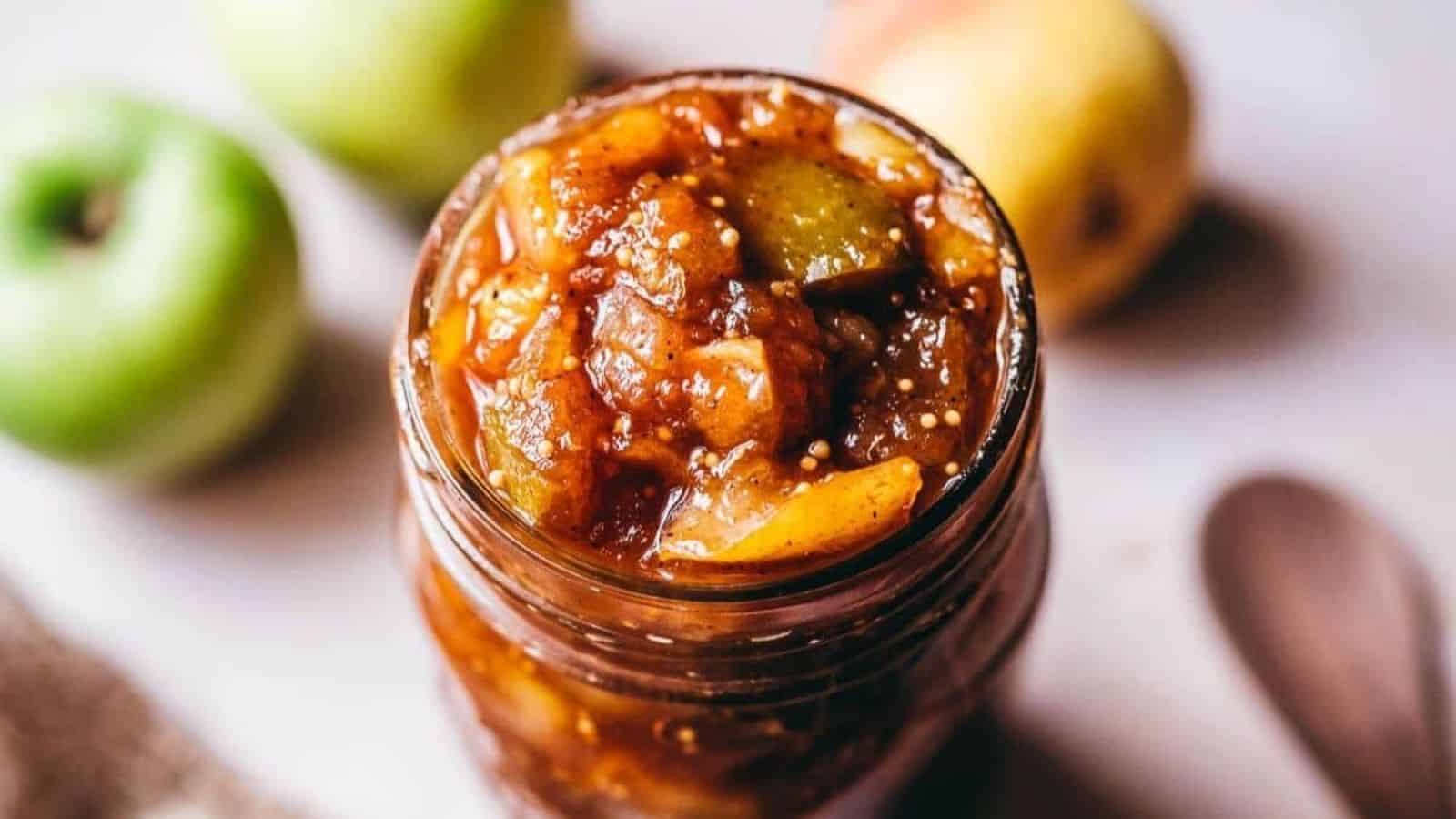 A glass jar filled with chunky apple chutney, with pieces of green apple visible, sits on a light surface next to fresh apples.